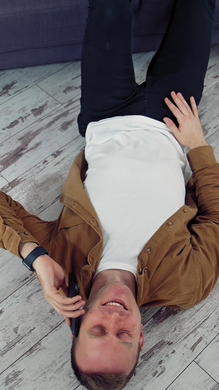 Young man talking the phone at home. Happy man having a conversation through the phone while lying on floor near the sofa. Top view. Quarantine. Vertical video