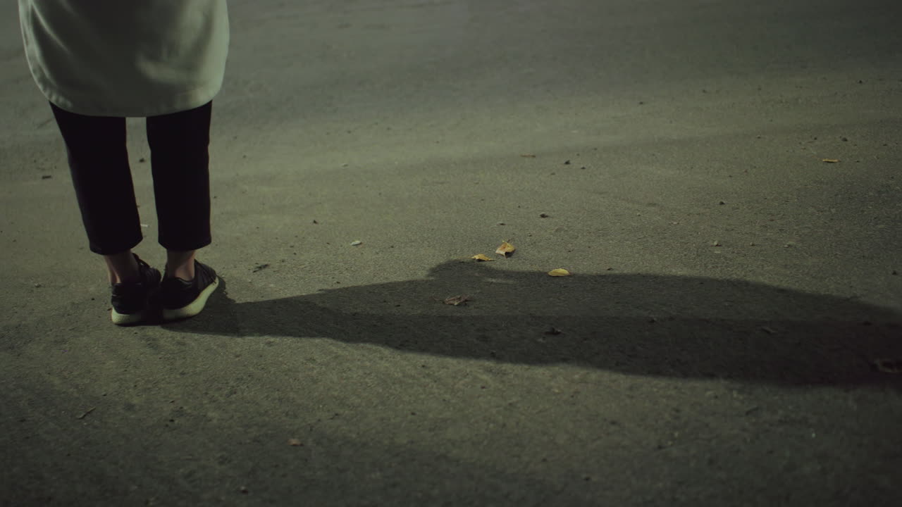 Leg view of woman in sneakers and coat standing quietly on roadside at night with long shadow cast on asphalt under artificial streetlight in urban setting, dry leaves scattered on the ground