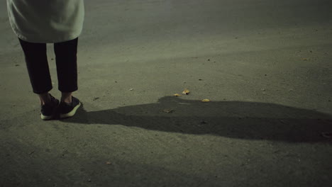 leg view of woman in sneakers and coat standing quietly on roadside at night with long shadow cast on asphalt under artificial streetlight in urban setting, dry leaves scattered on the ground
