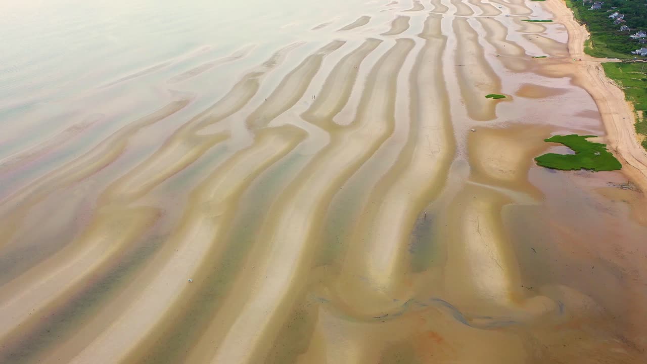 Wide aerial view of sandy shoreline at low tide where tide pools shimmer across rippled sand dunes, creating patterns of light and shadow under the soft glow of daylight