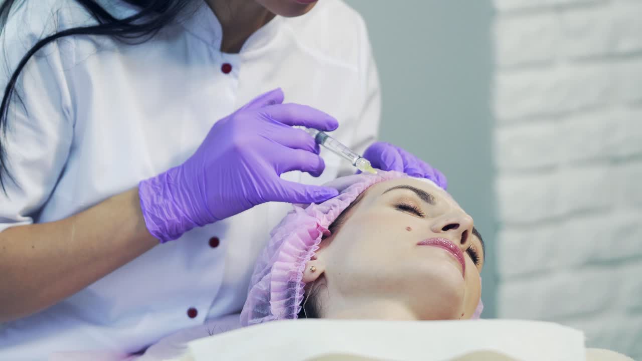 Rejuvenating facial procedure for tightening wrinkles of a young woman in clinic. Pretty doctor pricks girl's forehead