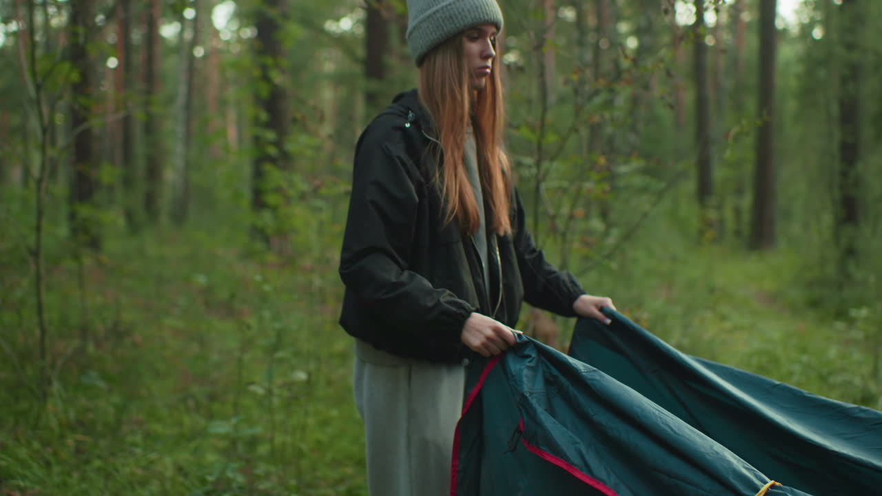 Woman in casual outfit picks up tent cover with gentle smile during outdoor camping in forest, lifting fabric while someone assists in background surrounded by trees and greenery