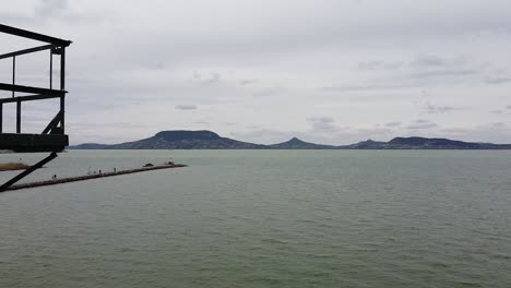 Vista-Aérea---Pasando-Por-Un-Pilar-De-Señal-De-Tormenta-En-El-Lago-Balaton-Con-Montañas-En-El-Fondo