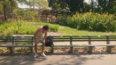 Medium-full-body-view-of-man-walking-up-to-park-bench-sitting-by-bag-and-relaxing,-space-for-text