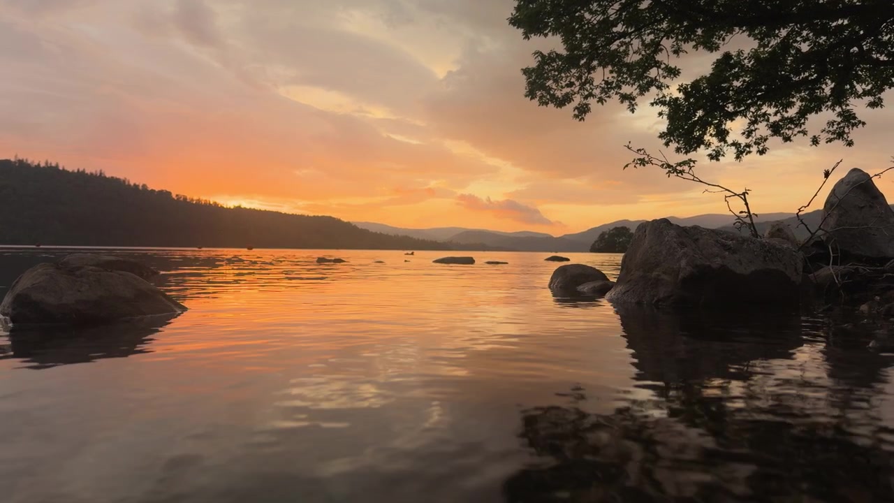 A captivating low-angle shot focusing on rocks in the foreground, with a wide view of Lake Windermere during a warm sunset, showcasing the vibrant colors reflecting on the water.