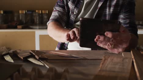 Midsection-of-caucasian-male-knife-maker-in-workshop,-using-tablet-and-smiling