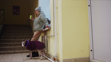 woman in grey beanie and green puffer jacket holding cup stands beside whippet in purple coat with toy around neck, both waiting in tiled indoor hallway near stairs in winter, casual urban moment