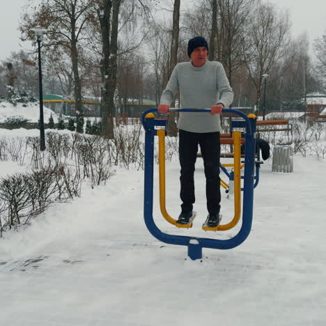 aged caucasian male stands on the simulator in winter outdoors. man exercising in the park on snowy day.