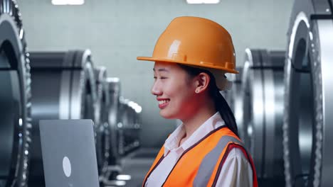 close up side view of asian female engineer with safety helmet working on a laptop and looking around while standing in metal factory