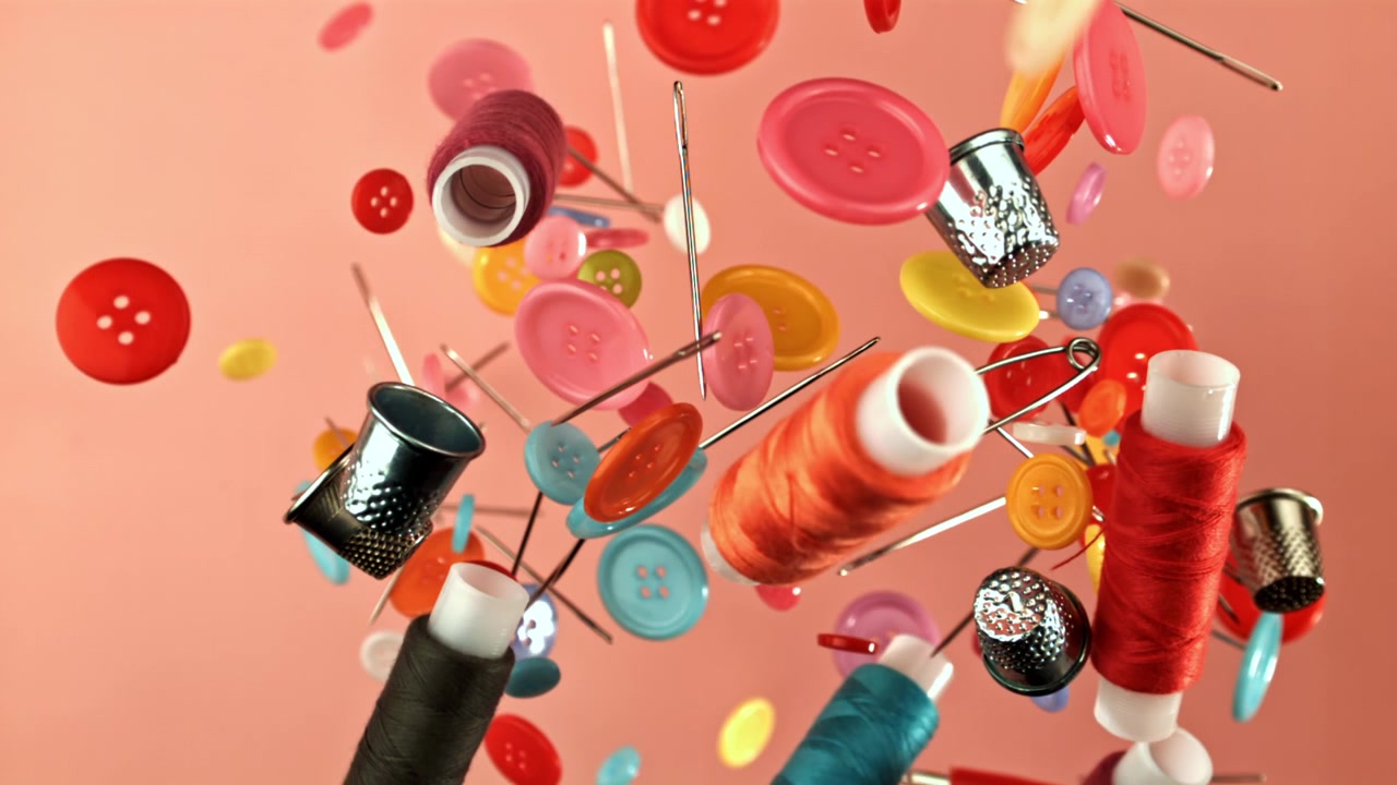 Bunte Nähknöpfe, Fäden und Fingerhüte fallen in Zeitlupe.
