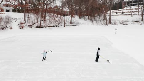 aerial, two kids clearing snow and ice skating on outdoor ice hockey rink on a frozen lake