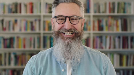 portrait of mature caucasian professor with beard laughing cheerful at camera in library bookshelf background wearing glasses