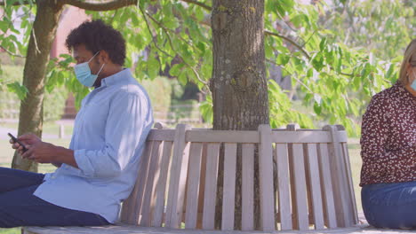 Socially-Distanced-Couple-In-Masks-Meet-In-Park-During-Health-Pandemic-Looking-At-Mobile-Phones