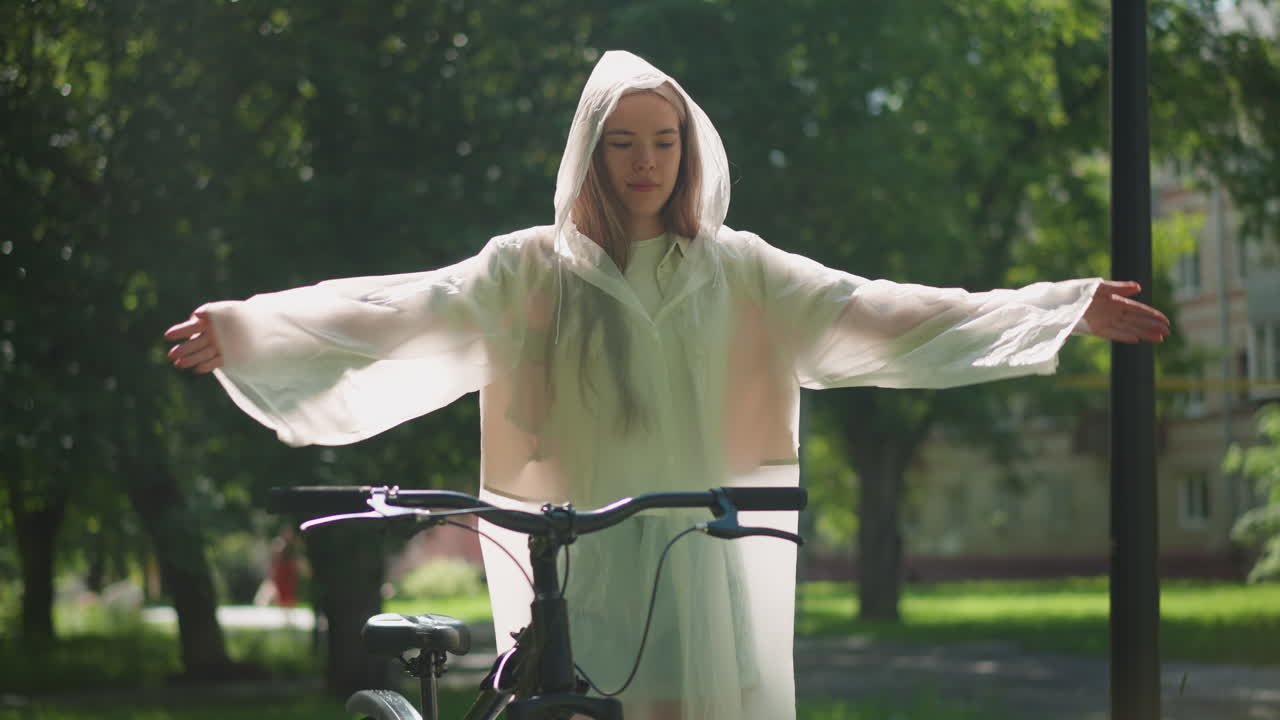 반투명한 레인코트를 입은 젊은 여성은 팔을 펴고 자전거 에 기게 서서 새의 날개처럼 손을 우아하게 흔들고, 빛은 잔잔한 초록색을 통해 뒷면에 건물을 필터링합니다.