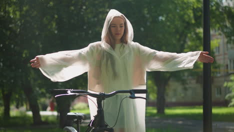 joven con impermeable translúcido alegremente está de pie junto a la bicicleta con los brazos extendidos, agitando con gracia sus manos como alas de pájaro, la luz del sol se filtra a través de la vegetación exuberante con el edificio en el fondo