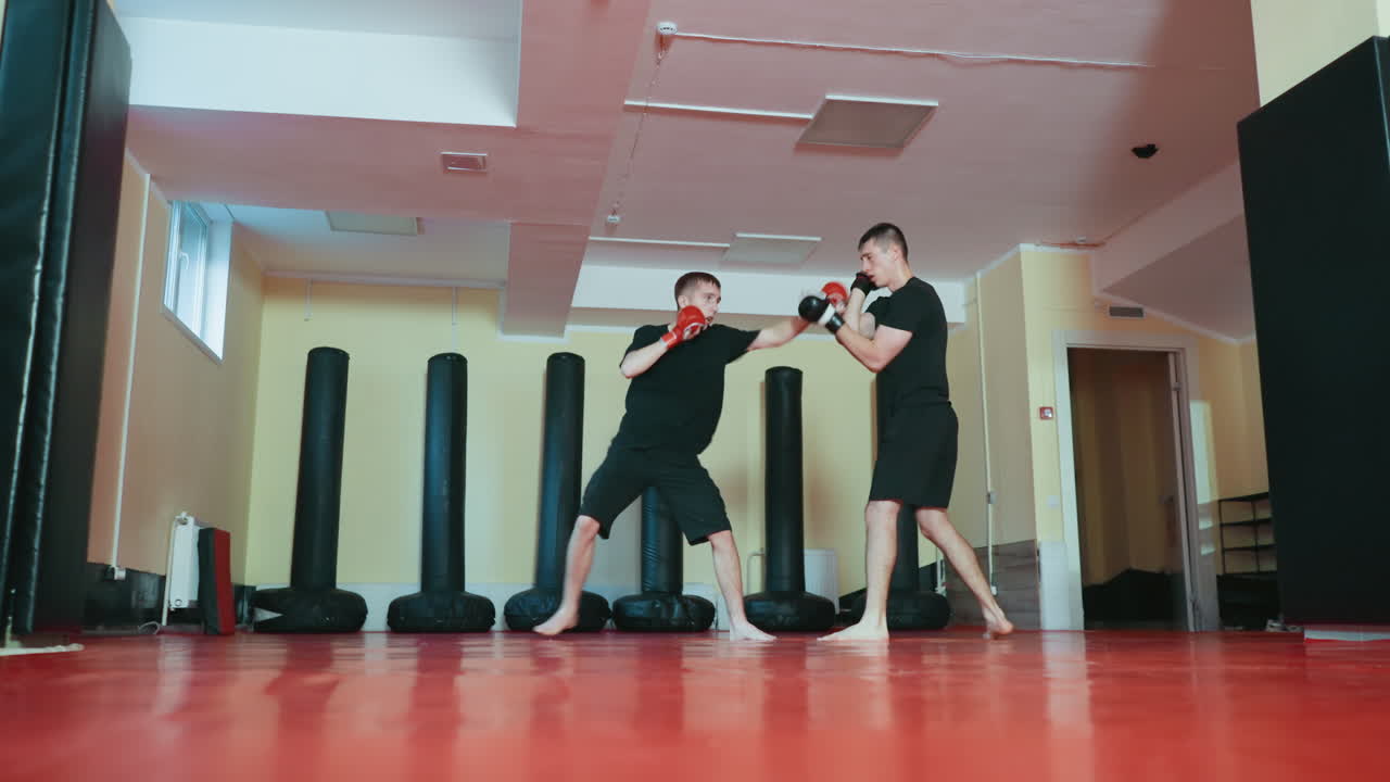 격투기 체육관 안에서 스파링 세션에 참여 중인 운동선수들의 모습이에요. 한 명이 펀치를 날리는 동안 상대는 방어하고 있으며, 둘 다 장갑을 끼고 붉은색 매트 위에서 검은색 운동복을 입고 샌드백으로 둘러싸여 있어요.