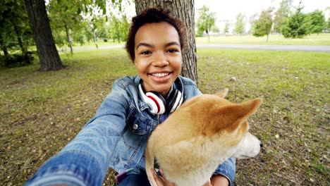 point of view shot of pretty african american girl taking selfie with cute puppy in city park holding camera, smiling and posing. modern technology and animals concept.