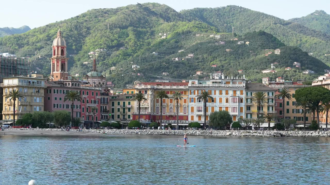 Ampia inquadratura diurna del colorato lungomare di Rapallo, Italia, con classici edifici liguri, rigogliose colline verdi e persone che fanno paddleboard su acqua calma. Una costa urbana vibrante, scenografica e invitante.
