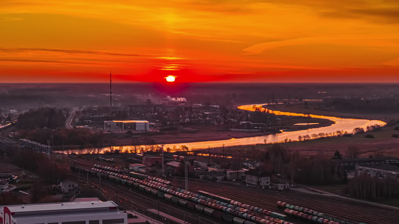 A Fiery Sunset Bathes the River and Rail Yard in Warm Orange Light as it Winds Through the Industrial Town - Timelapse
