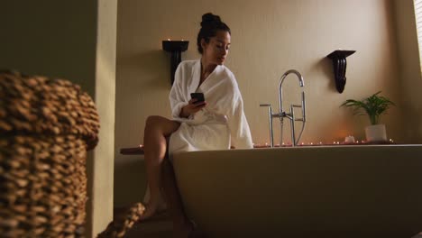 Biracial-woman-sitting-in-robe-on-bathtube,-preparing-bath,-using-smartphone