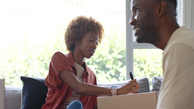 Diverse couple at home, woman writing box label with marker while man watching, preparing move