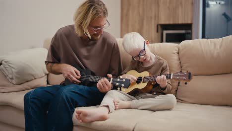 A-little-albino-boy-with-white-hair-in-blue-glasses-together-with-his-dad,-a-blond-man-in-glasses-with-a-beard-learns-to-play-the-ukulele-while-sitting-on-a-cream-colored-large-sofa-in-a-modern-apartment-in-the-evening