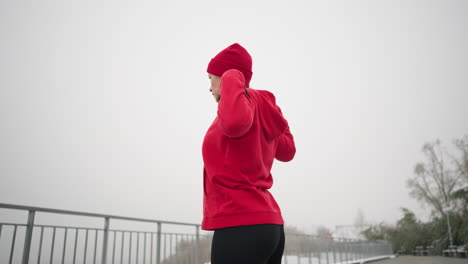 vista lateral de una mujer realizando un estiramiento de la mano girando el brazo al aire libre en una rutina de fitness, vestida con ropa atlética, suelo cubierto de nieve, árboles helados, banco, barandilla de hierro en un parque sereno