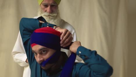 Fotografía-De-Estudio-De-Un-Hombre-Sikh-Mayor-Que-Ayuda-A-Un-Hombre-Sikh-Más-Joven-A-Atar-La-Tela-Para-Un-Turbante-Contra-Un-Fondo-Liso,-Filmada-En-Tiempo-Real-1