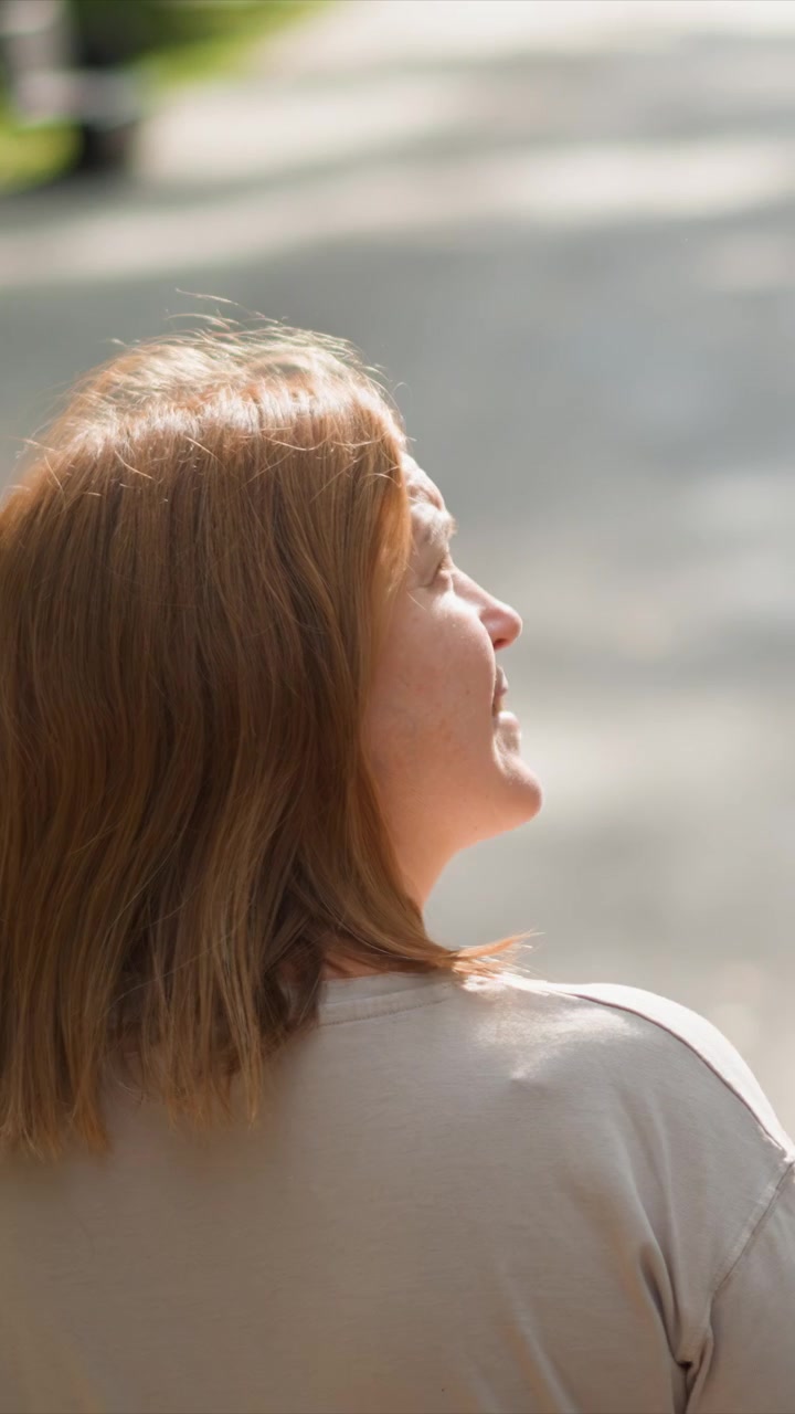 Il soggetto ha capelli castano-rossastri lunghi fino alle spalle. L'abbigliamento di colore chiaro contrasta con la luce solare intensa. Ambiente esterno luminoso con ombre morbide su capelli e collo.