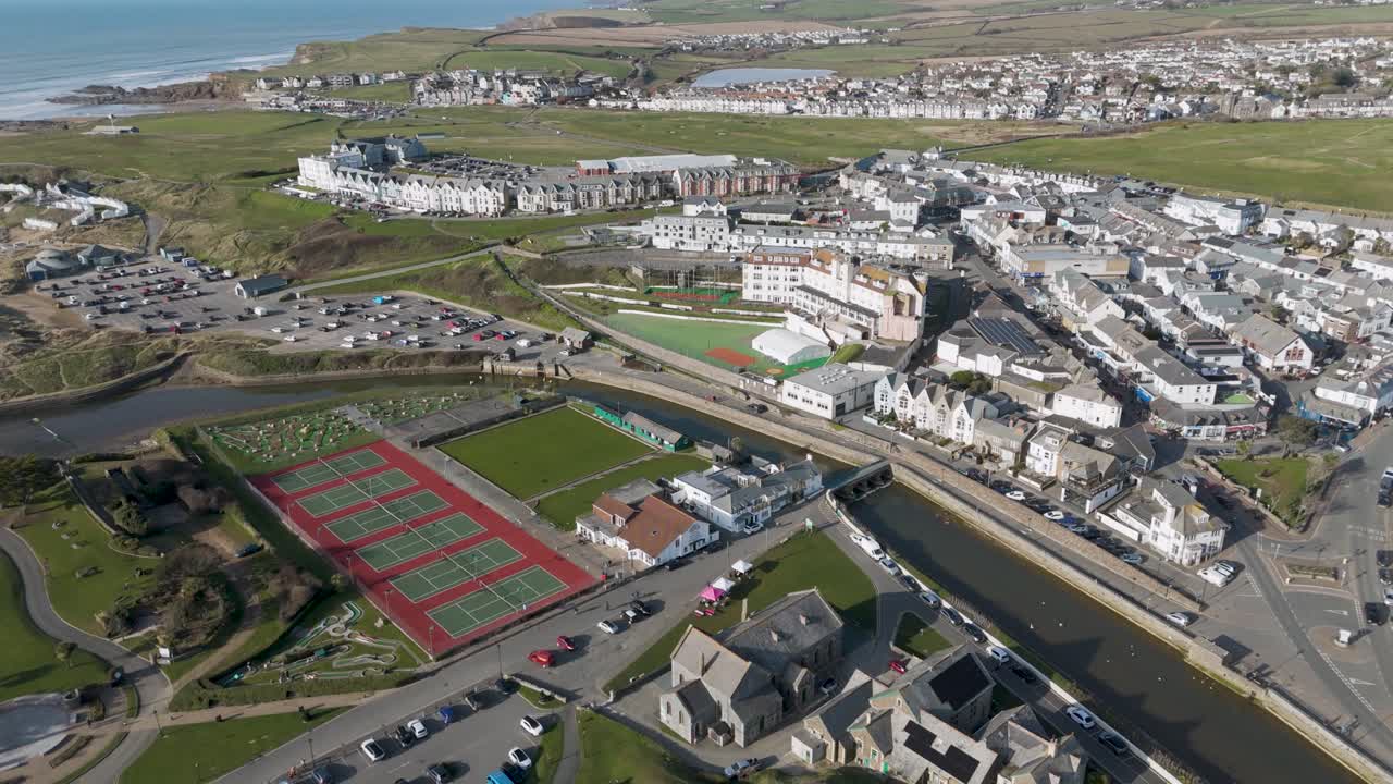 High aerial of Bude seaside town with white houses climbing coastal slope above beach, bordered by tennis courts, playing fields and dramatic Atlantic cliffs