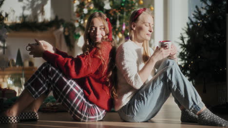 happy sisters enjoying coffee while sitting on floor at home during christmas