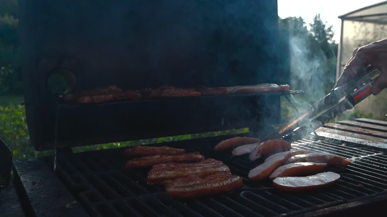 Golden brown and red meat, ribs and sausages being cooked on a smoking hot grill with a mans hand turning the sausages around in slow motion. Bokeh blurry green sunny garden in the background