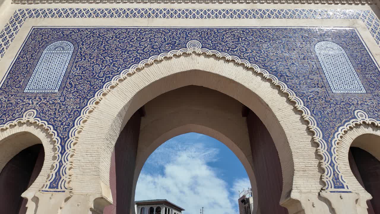 Traditional moroccan architecture of Bab Bou Jeloud gate with blue mosaic tiles and white walls framing the blue sky with clouds