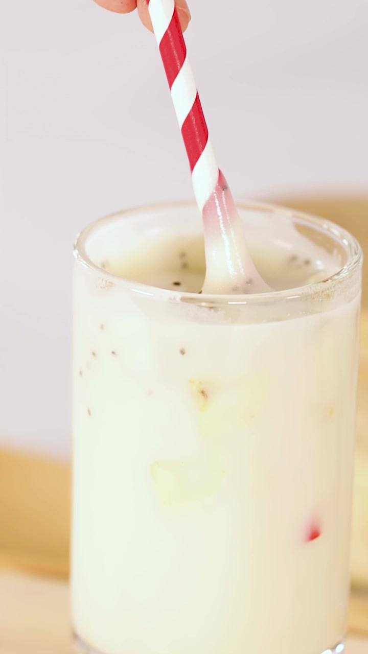 Mão mexe milk-shake de banana em copo transparente usando canudo listrado, iluminação brilhante, close-up.