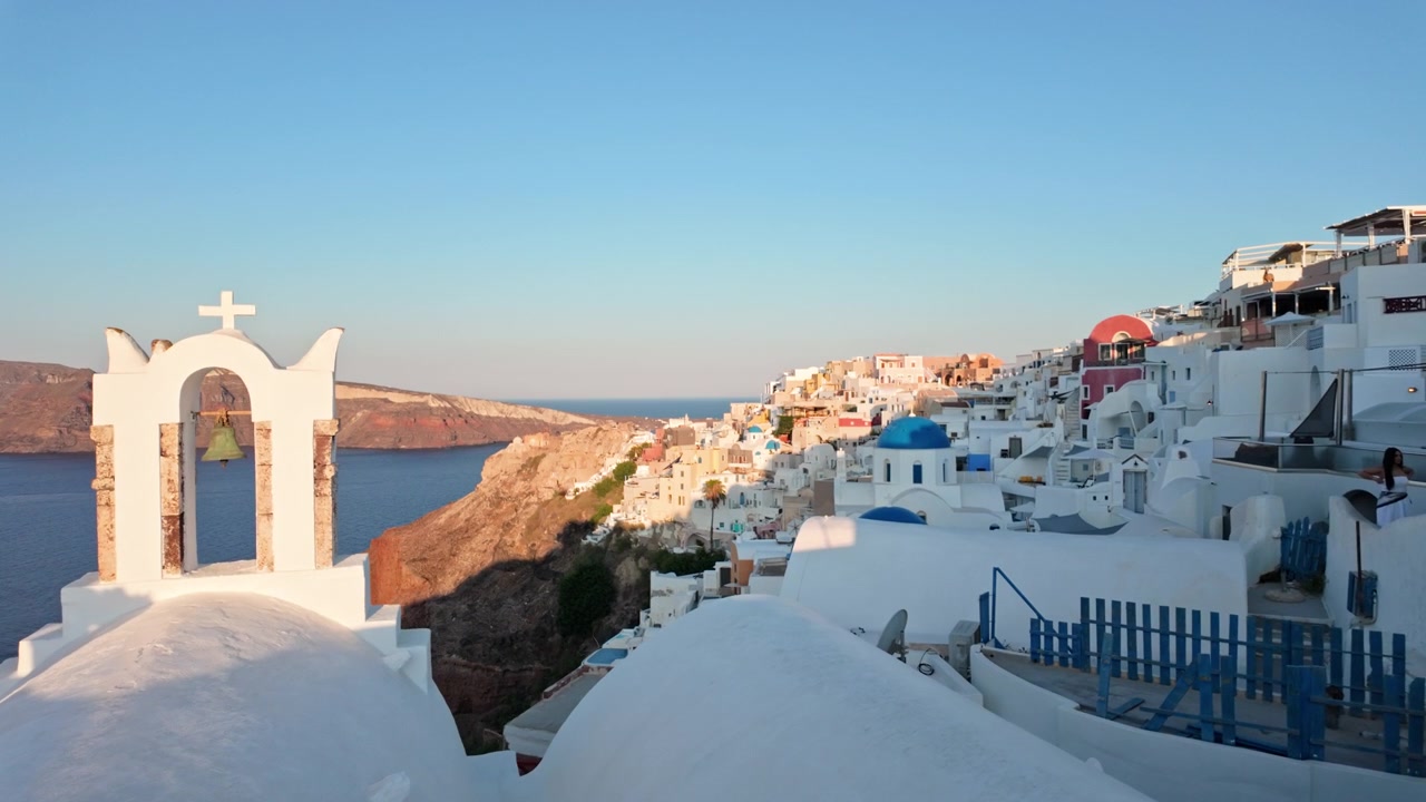 Torre sineira da igreja e cúpulas brancas dominam uma vila na encosta e o mar calmo sob a luz quente do pôr do sol, vista clássica do horizonte de Santorini