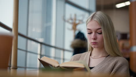 back view of student preparing for exam, focused as she flips to a new page of her textbook while sitting at a table in a study area with a cup of coffee and phone beside him