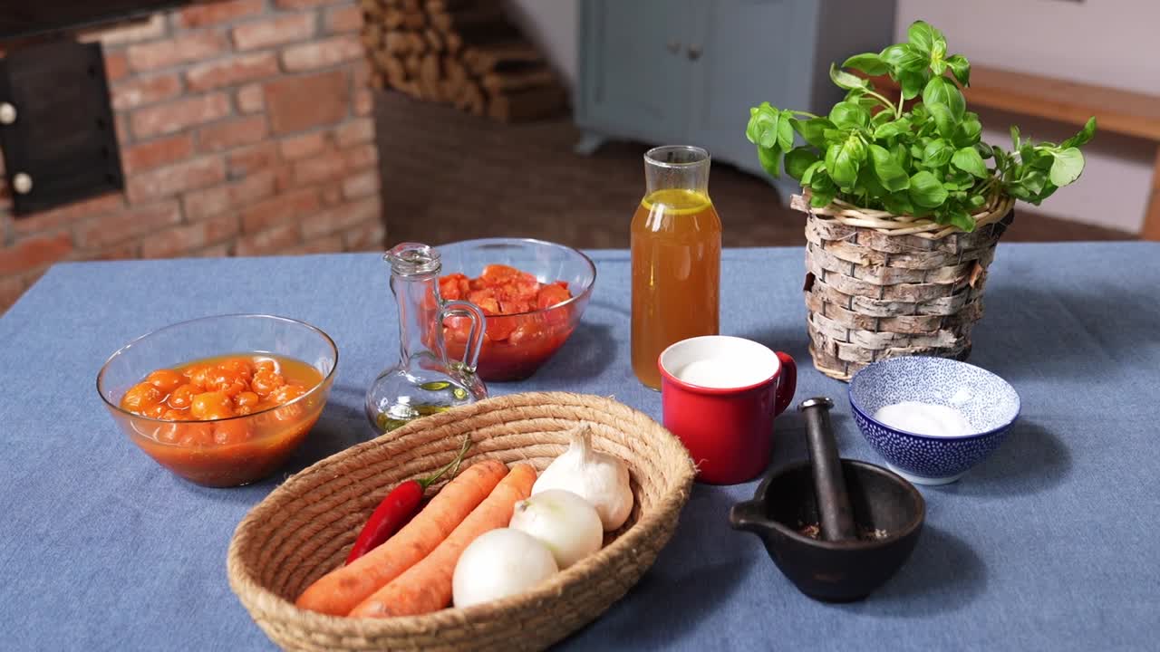A colorful arrangement of fresh ingredients prepared for cooking tomato soup. The table displays carrots, onions, garlic, chili, tomatoes, oil, vinegar, herbs, salt, and broth on a rustic blue cloth