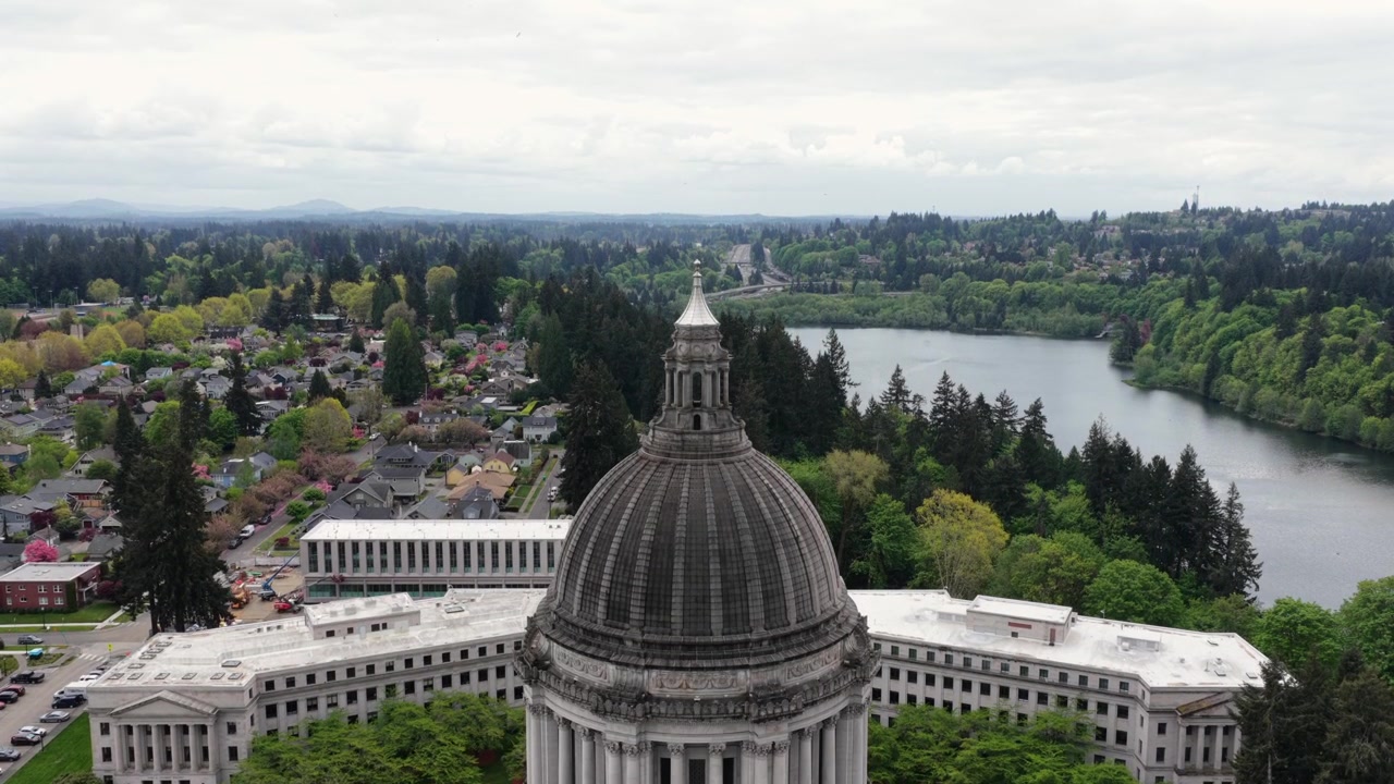 Cupola del Campidoglio dello Stato di Washington a Olympia con il campus legislativo e il Lago Capitol, sullo sfondo un tranquillo quartiere residenziale e alberi verdi lussureggianti sotto un cielo coperto negli USA. Ampia inquadratura aerea ascendente.