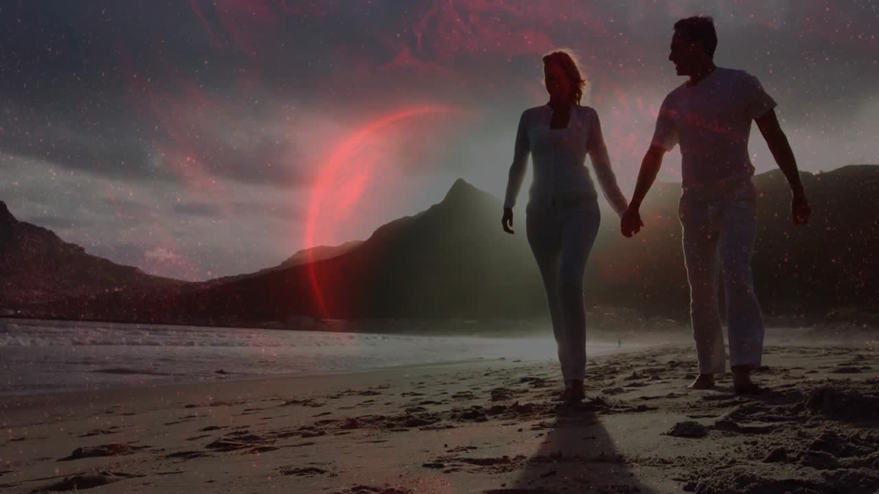 Una coppia che cammina dalla riva, si avvicina tenendosi per mano per un viaggio, con un bagliore rosso su una cresta dietro di loro.