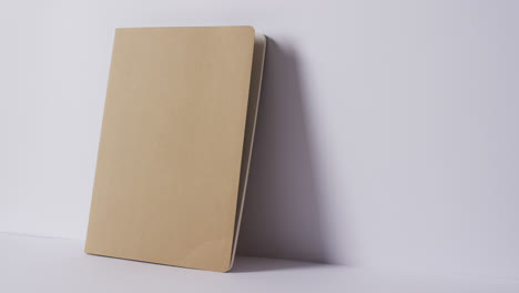 Close-up-of-closed-beige-book-leaning-on-wall-with-copy-space-on-white-background-in-slow-motion
