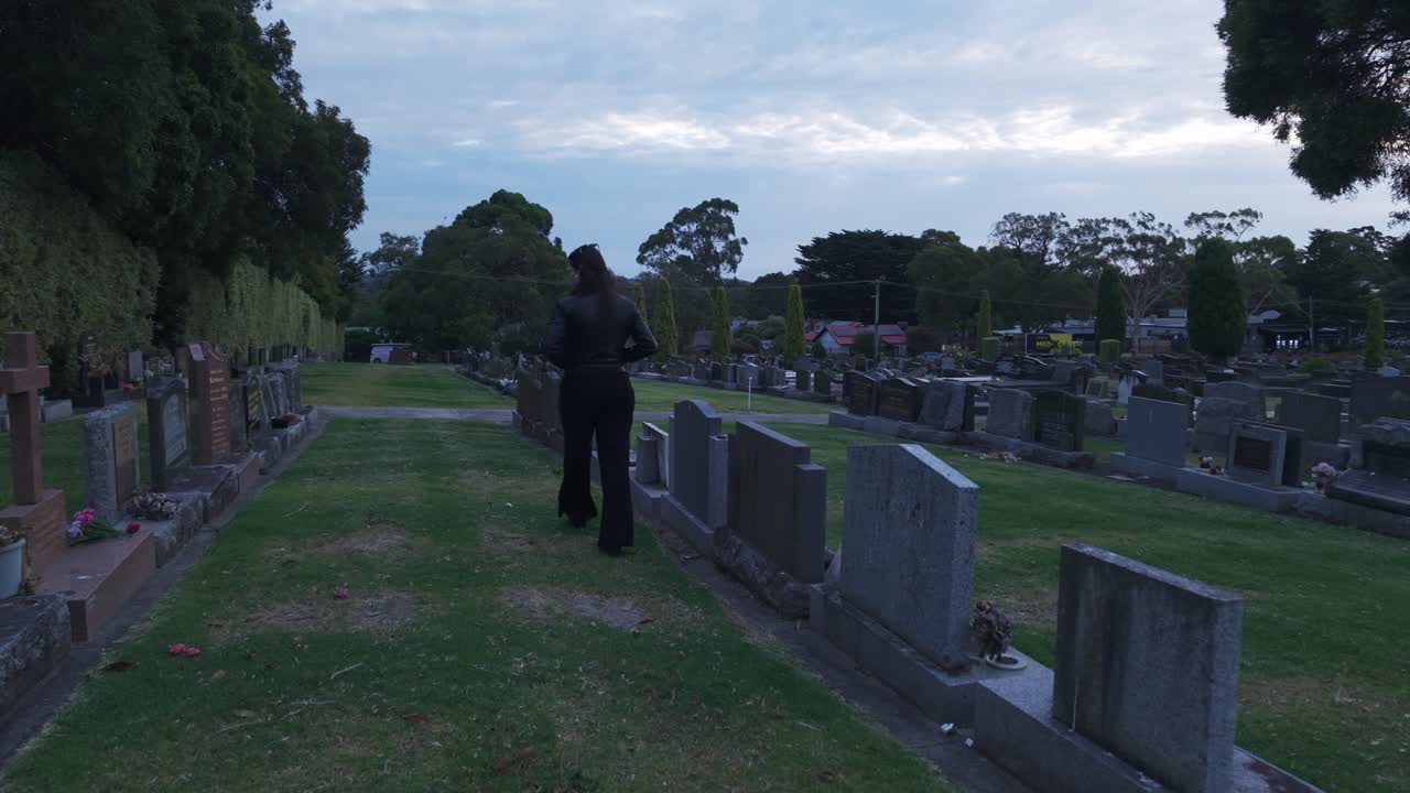 Una ragazza ripresa di spalle che cammina attraverso un tranquillo cimitero al tramonto a Ferntree Gully, Melbourne. La scena è serena, con illuminazione soffusa e una prospettiva dinamica.