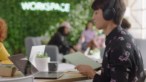 young caucasian man student using laptop computer working on creative project browsing online research writing notes enjoying study listening to music wearing headphones in trendy office workspace