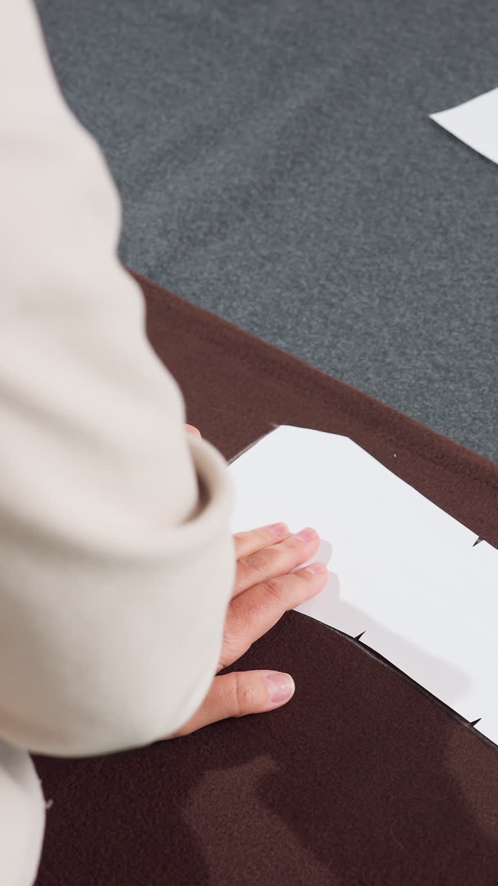 hands tracing fabric pattern on table, bespoke tailor aligns paper template on brown cloth, presses edge with palm for smooth adhesion, pins guide marks for cutting, pattern cutter prepares layout
