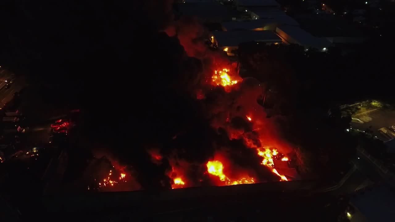 Night aerial view of a fierce warehouse fire, captured by a drone. Intense flames and thick smoke light up the night as firefighters struggle to control the blaze