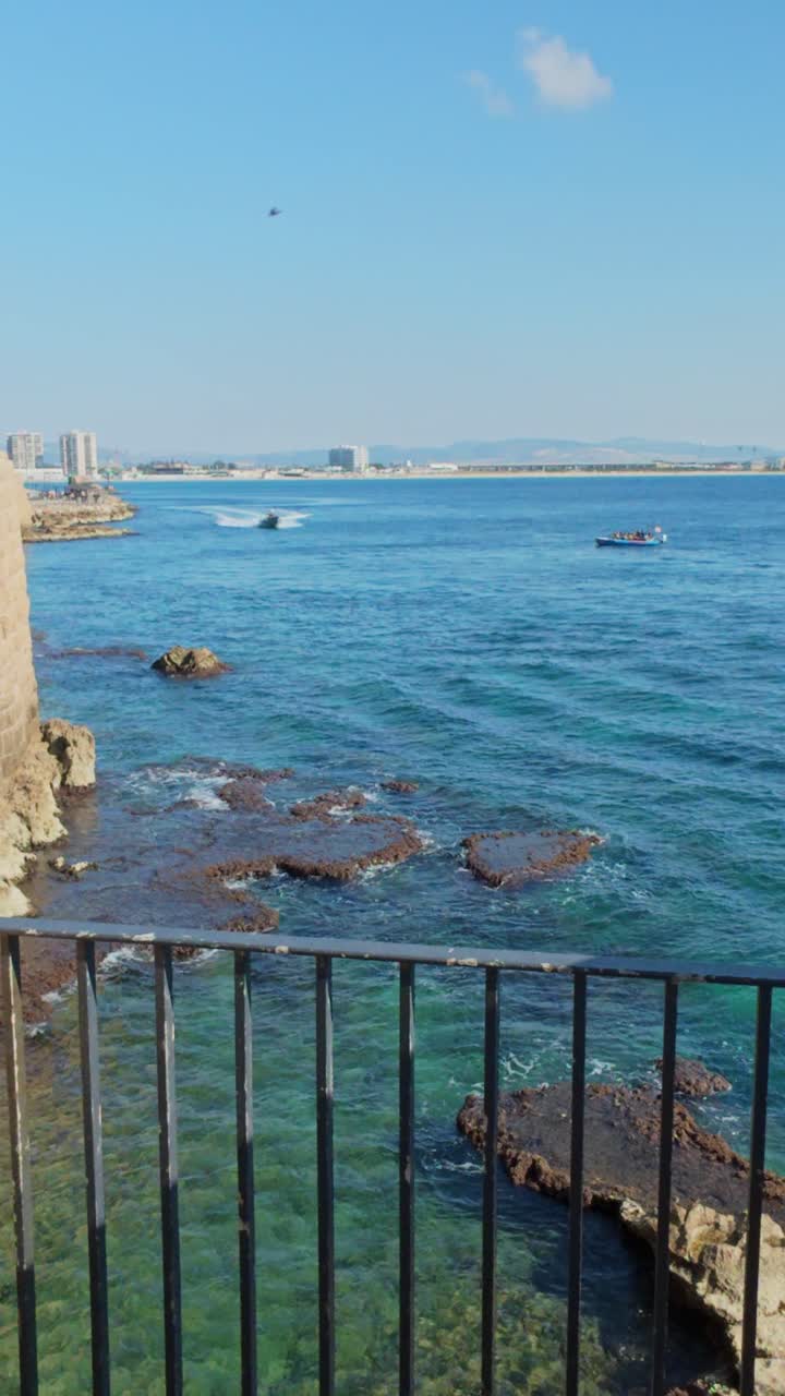 Guardando attraverso una recinzione di metallo nero una barca che passa sopra il fondale roccioso ad Akko, Israele.