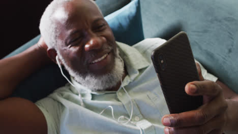 hombre afroamericano usando un teléfono inteligente mientras está tendido en el sofá en casa
