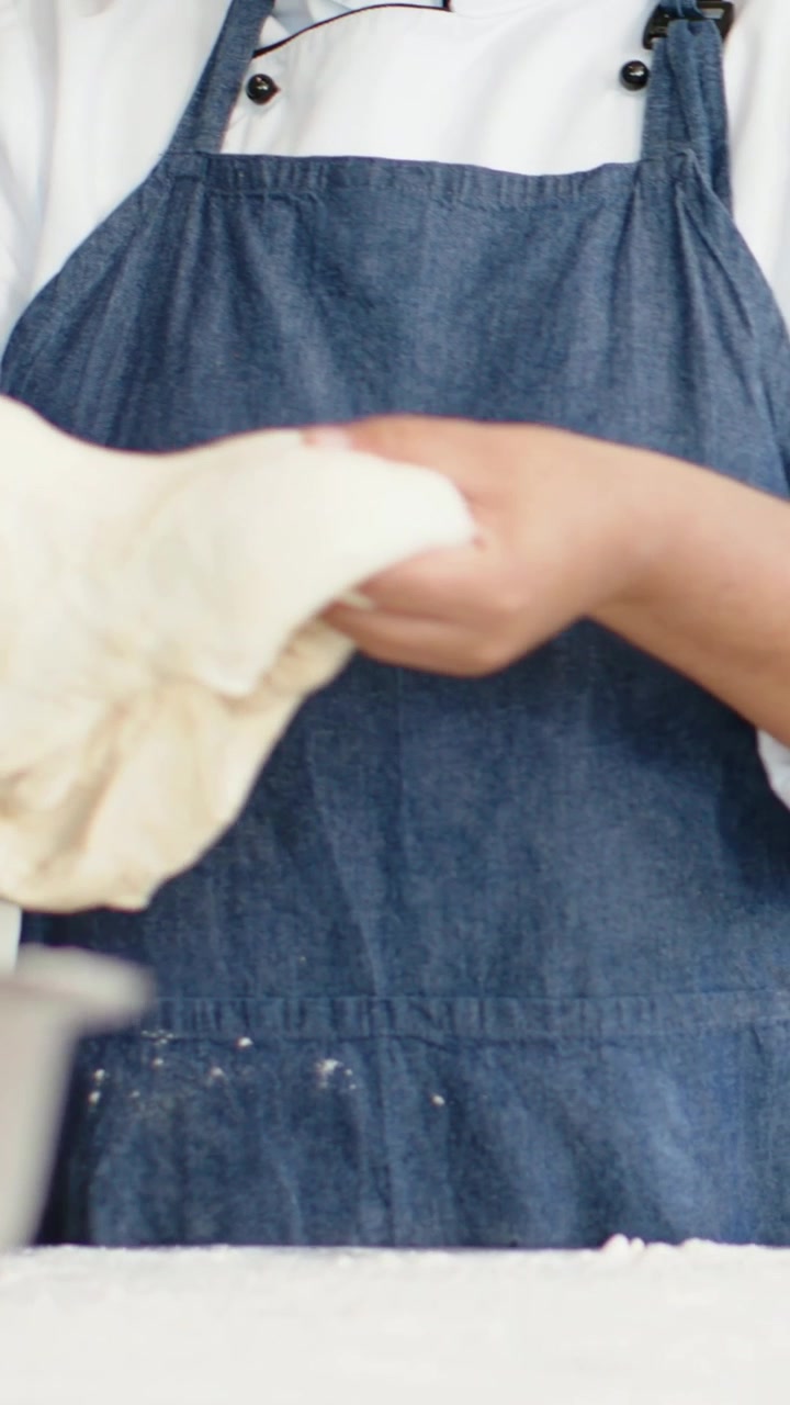Video verticale: Chef in grembiule che impasta con le mani in una ciotola sul bancone, preparando l'impasto e tenendo uno straccio.