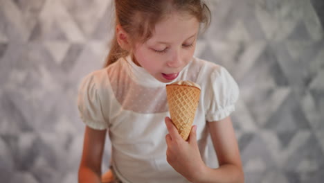 little girl in white top holding ice cream cone, looking down with sweet smile, standing against patterned gray wall, capturing pure joy and innocence of childhood