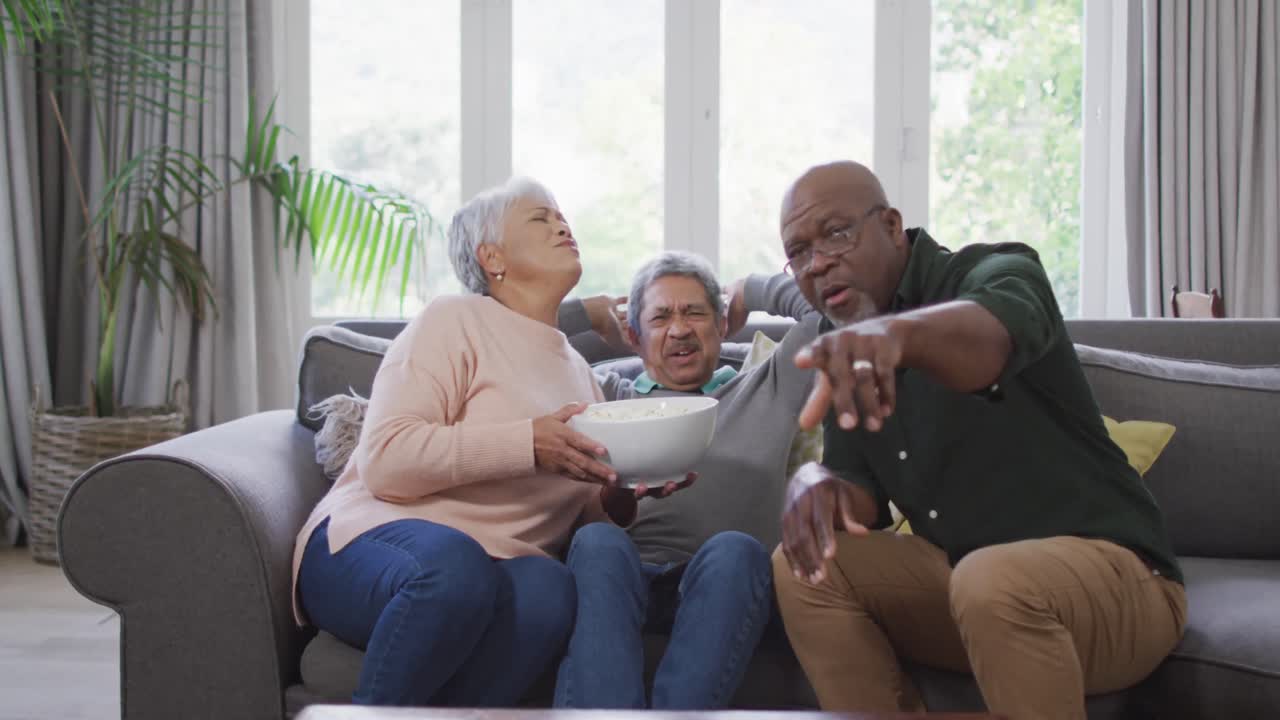 Happy biracial senior couple and african american friend watching tv with popcorn