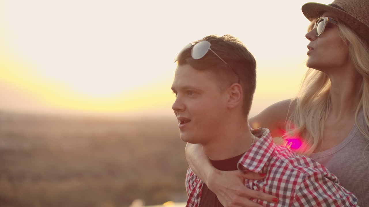 Young couple moves and spins in a dance at sunset. Women with blond hair in a hat and glasses is sitting on her boyfriend's back.